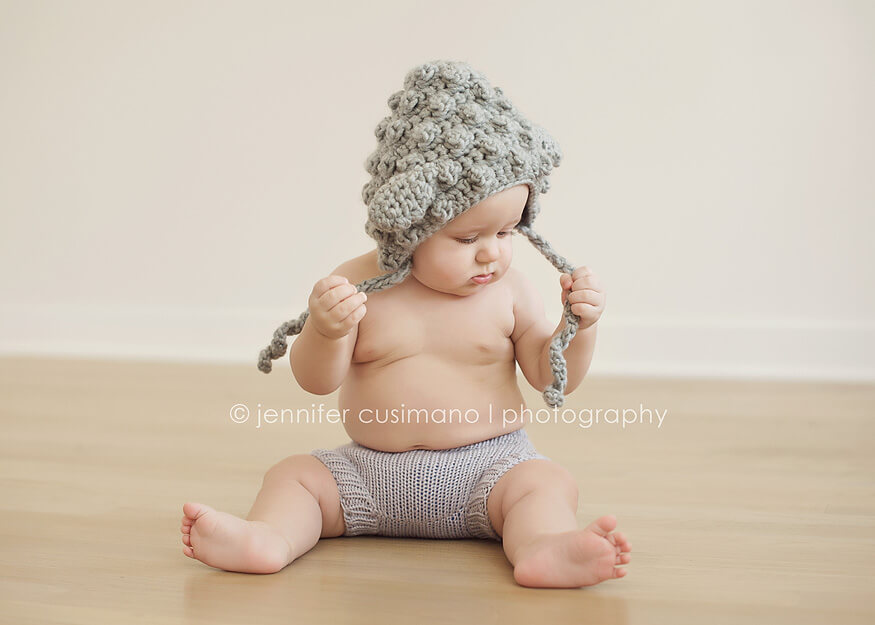 baby boy sitting on hard wood floors wearing a grey little lamb hat