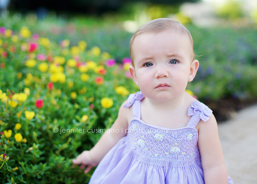 9 month old baby girl is purple dress sitting in front of yellow and pink flowers